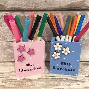 May include: Two square wooden pencil holders, one light pink with pink star accents and the text Miss Edmondson, and one light blue with white flower accents and the text Miss Wareham, both filled with an assortment of colorful felt-tip markers.