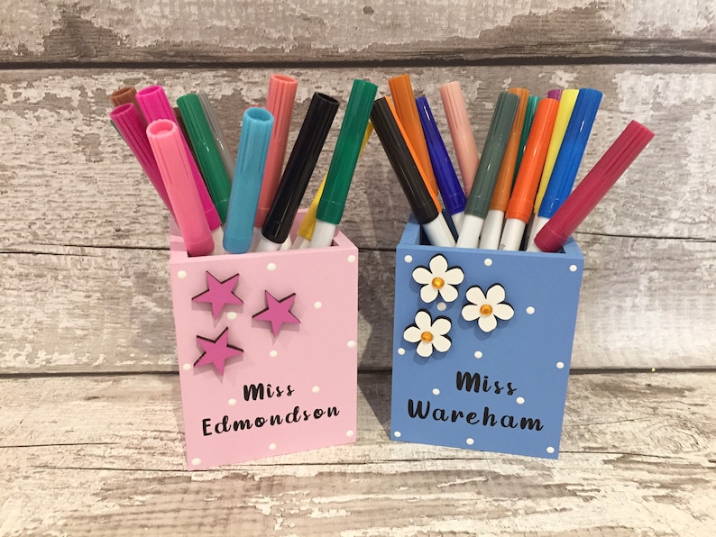 May include: Two square wooden pencil holders, one light pink with pink star accents and the text Miss Edmondson, and one light blue with white flower accents and the text Miss Wareham, both filled with an assortment of colorful felt-tip markers.