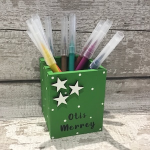 May include: A square green wooden pencil holder featuring white polka dots and three white star cutouts on the front. The name Otis Merrey is printed in black text on the lower front. Several colorful markers with clear caps are stored inside.
