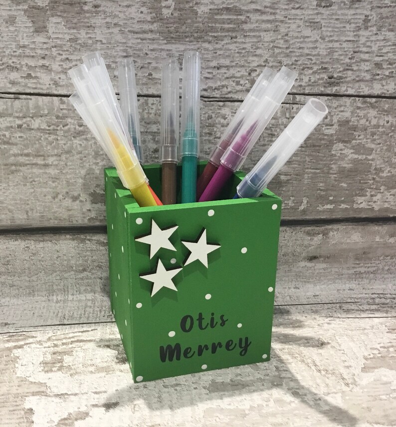 May include: A square green wooden pencil holder featuring white polka dots and three white star cutouts on the front. The name Otis Merrey is printed in black text on the lower front. Several colorful markers with clear caps are stored inside.