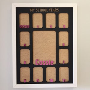 May include: A white-framed photo collage board titled MY SCHOOL YEARS in gold lettering. It features a black mat with twelve rectangular openings for photos, labeled with pink numbers from Reception to 11, and the name Cassie in pink.