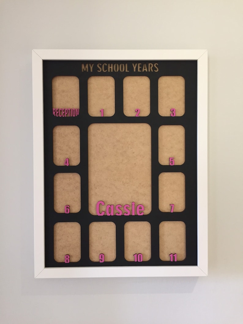 May include: A white-framed photo collage board titled MY SCHOOL YEARS in gold lettering. It features a black mat with twelve rectangular openings for photos, labeled with pink numbers from Reception to 11, and the name Cassie in pink.