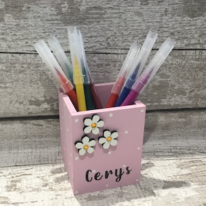 May include: A light pink wooden pencil holder with white polka dots, decorated with three white flower embellishments featuring gold centers. The name Cerys is printed in black script on the front, and it contains several colorful markers.