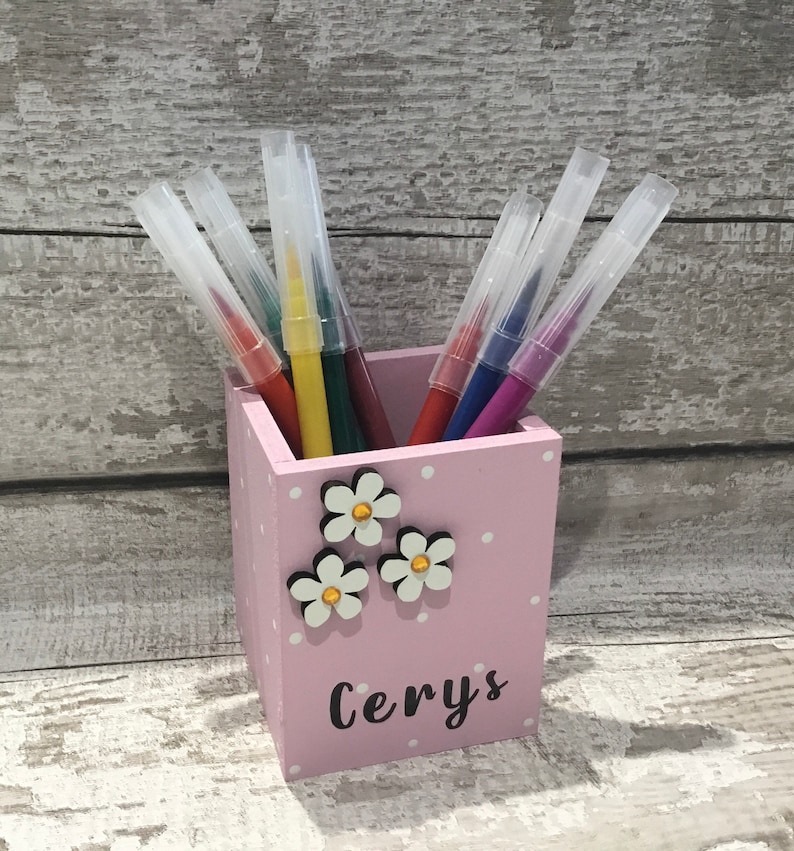 May include: A light pink wooden pencil holder with white polka dots, decorated with three white flower embellishments featuring gold centers. The name Cerys is printed in black script on the front, and it contains several colorful markers.