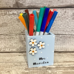 May include: A light blue square pen holder with white polka dots, three white flower embellishments with yellow centers, and the black text Mrs Marsden printed on the front, filled with an assortment of colorful felt-tip markers.