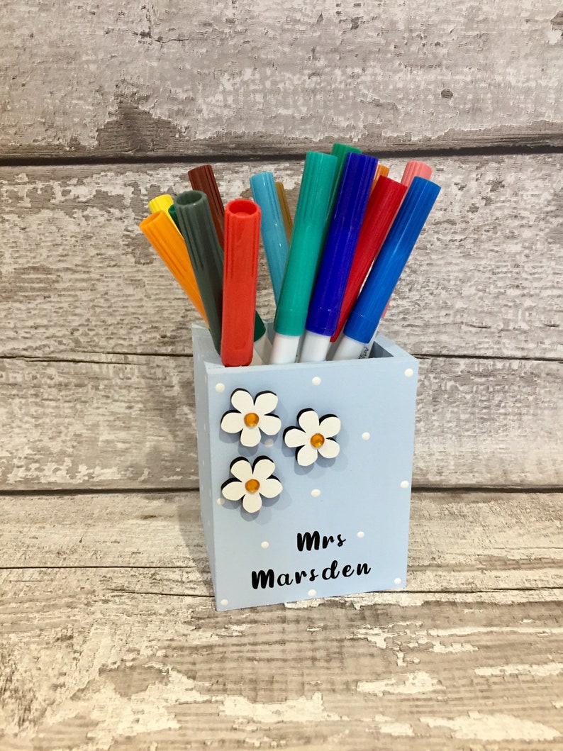 May include: A light blue square pen holder with white polka dots, three white flower embellishments with yellow centers, and the black text Mrs Marsden printed on the front, filled with an assortment of colorful felt-tip markers.