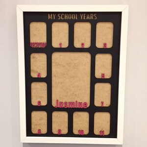 May include: A white framed collage picture frame with a black mat board titled MY SCHOOL YEARS in gold. It features twelve small rectangular openings labeled with pink text for Reception through 11, and a large central opening labeled Jasmine.