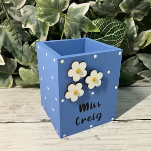 May include: A square blue wooden pencil holder featuring a white polka dot pattern, three white flower embellishments with yellow centers, and the black text "Miss Creig" printed on the front side of the container for desk organization.