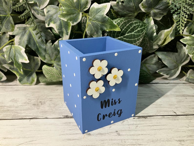 May include: A square blue wooden pencil holder featuring a white polka dot pattern, three white flower embellishments with yellow centers, and the black text "Miss Creig" printed on the front side of the container for desk organization.