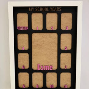 May include: A white rectangular picture frame with a black mat board titled MY SCHOOL YEARS. It features twelve small photo slots labeled Reception through 11 in pink, and a larger central slot with the name Esme in pink lettering.
