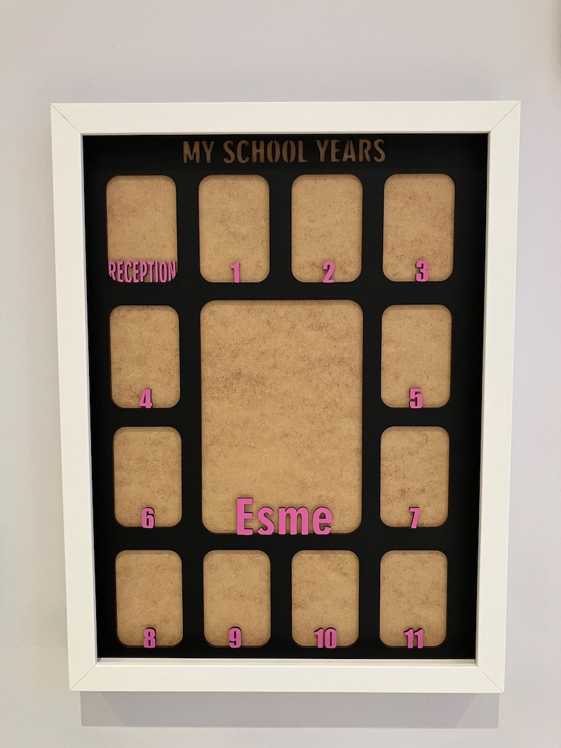 May include: A white rectangular picture frame with a black mat board titled MY SCHOOL YEARS. It features twelve small photo slots labeled Reception through 11 in pink, and a larger central slot with the name Esme in pink lettering.