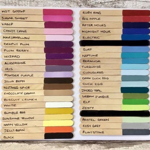 May include: A color chart featuring two columns of wooden craft sticks, each tipped with a unique paint color and labeled with names like Hot Gossip, Ruby Ring, and Black. The sticks are arranged in rows, displaying a wide spectrum of shades.