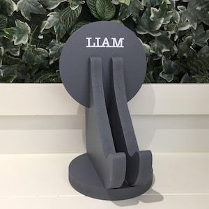 K&ouml;nnte beinhalten: Grauer Holz-Telefonst&auml;nder mit einer runden Oberseite, auf der "LIAM" in wei&szlig;en Buchstaben steht. Der St&auml;nder ist in Form einer sitzenden Person gestaltet.