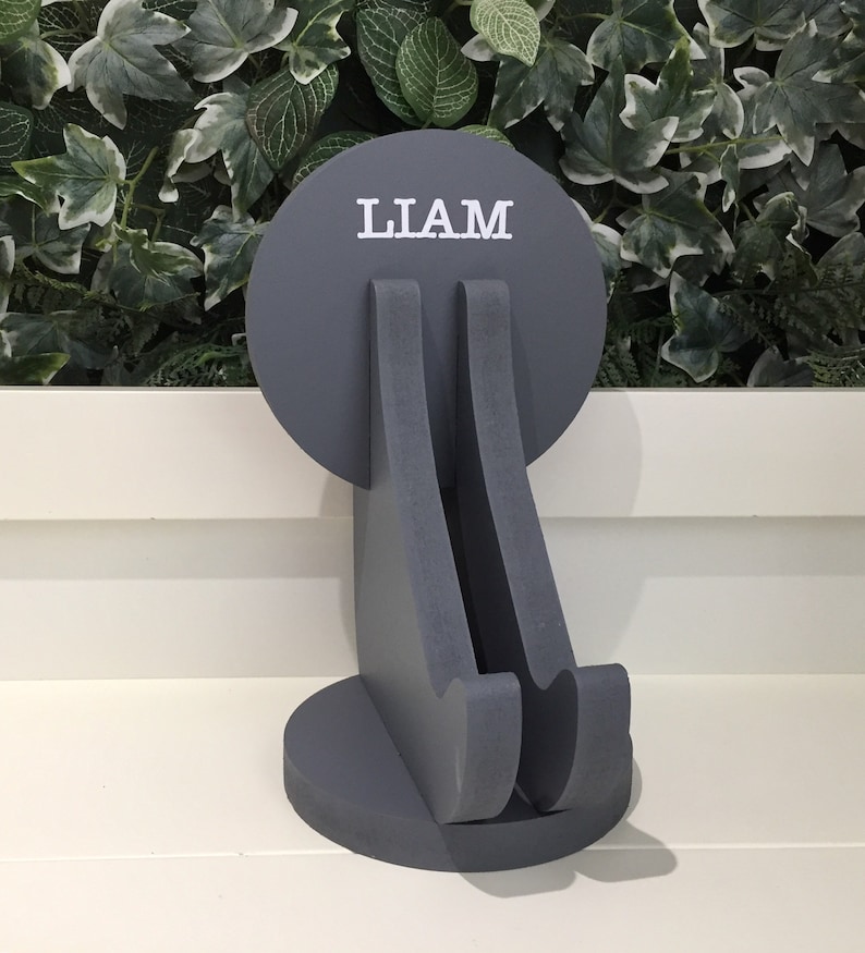 K&ouml;nnte beinhalten: Grauer Holz-Telefonst&auml;nder mit einer runden Oberseite, auf der "LIAM" in wei&szlig;en Buchstaben steht. Der St&auml;nder ist in Form einer sitzenden Person gestaltet.