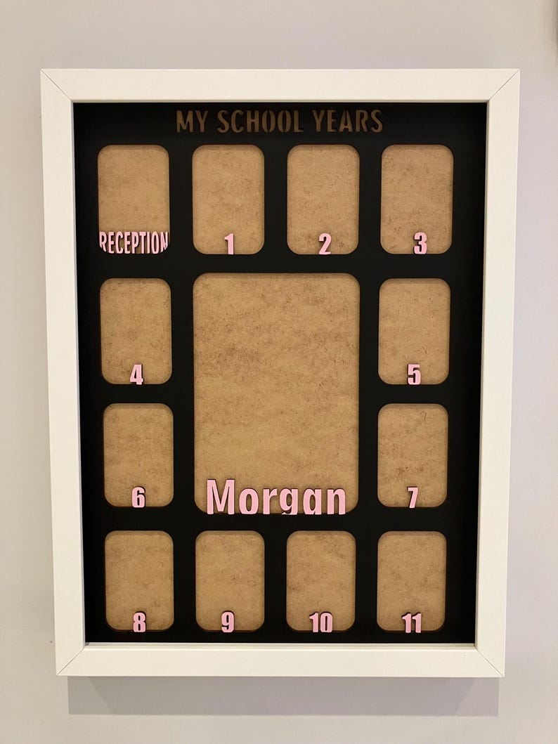 May include: A white rectangular frame containing a black mat with the text MY SCHOOL YEARS at the top. It features twelve small photo slots labeled Reception through 11 in pink, and a larger central slot with the name Morgan in pink lettering.
