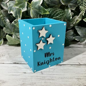 May include: A square turquoise wooden pencil holder featuring white polka dots, three small white star cutouts, and the black text "Mrs Knighton" printed on the front side. The container has an open top and a flat base for stability.
