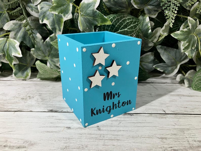May include: A square turquoise wooden pencil holder featuring white polka dots, three small white star cutouts, and the black text "Mrs Knighton" printed on the front side. The container has an open top and a flat base for stability.