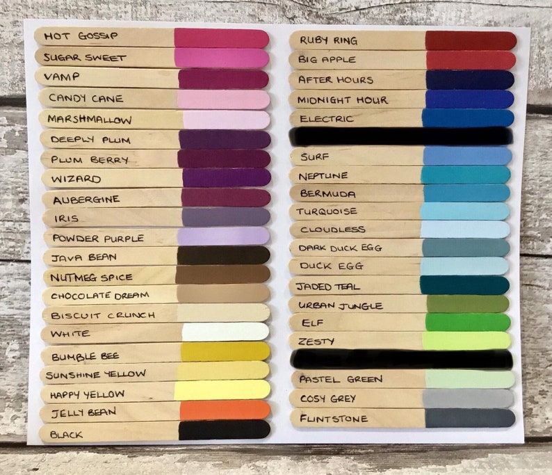 May include: A color chart featuring two columns of wooden craft sticks, each painted with a distinct color swatch at one end and labeled with names like Hot Gossip, Ruby Ring, Black, and Flintstone in black ink on the natural wood surface.