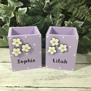 May include: Two light purple wooden pencil holders with a square shape. Each holder features white polka dots, three white flower cutouts with yellow centers, and a personalized name in black script, specifically the names Sophie and Lilah.