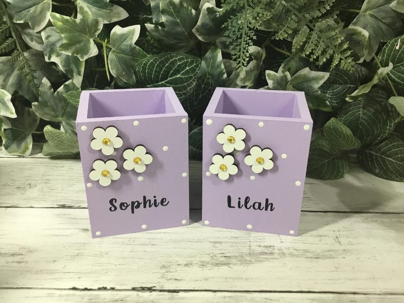 May include: Two light purple wooden pencil holders with a square shape. Each holder features white polka dots, three white flower cutouts with yellow centers, and a personalized name in black script, specifically the names Sophie and Lilah.