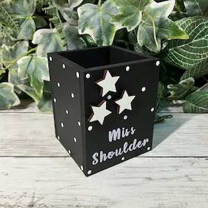 May include: A black square pencil holder featuring white polka dots, three white star cutouts, and the white text "Miss Shoulder" printed on the front. The container has an open top and a flat base, designed for holding stationery items.