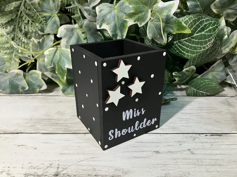 May include: A black square pencil holder featuring white polka dots, three white star cutouts, and the white text "Miss Shoulder" printed on the front. The container has an open top and a flat base, designed for holding stationery items.