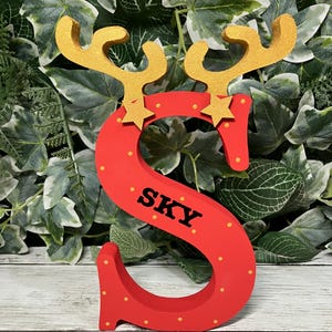 May include: Red wooden letter S with gold reindeer antlers and yellow stars. The letter is decorated with yellow dots and the name "SKY" is written in black letters.