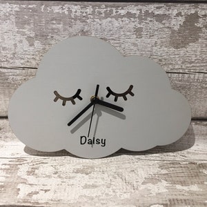 May include: A grey wooden cloud-shaped clock with black hands and eyelashes. The clock has the name "Daisy" written below the clock face.