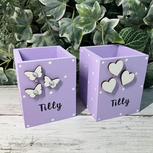 May include: Two square light purple wooden pencil holders featuring white polka dots and the name Tilly printed in black. One holder is decorated with three white butterfly cutouts, while the other features three white heart-shaped wooden cutouts.