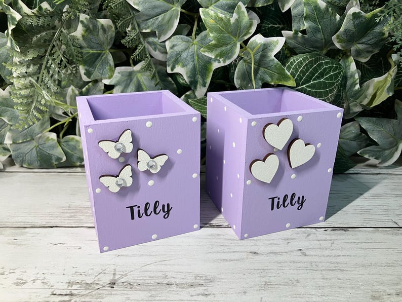 May include: Two square light purple wooden pencil holders featuring white polka dots and the name Tilly printed in black. One holder is decorated with three white butterfly cutouts, while the other features three white heart-shaped wooden cutouts.