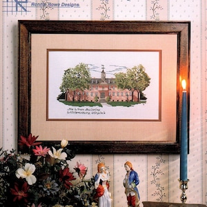 May include: A framed cross-stitch pattern of the Wren Building in Williamsburg, Virginia. The building is a light brown color with white trim and a green roof. The cross-stitch is surrounded by a brown frame and is displayed on a table with a floral arrangement and two figurines.