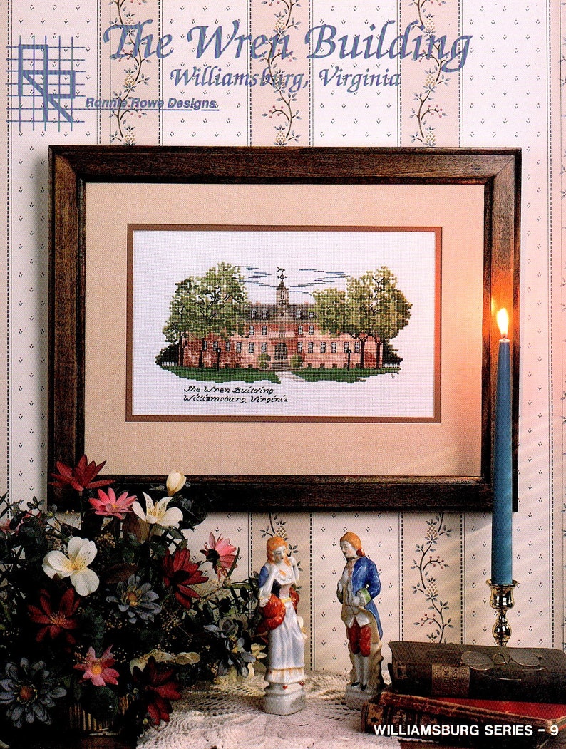 May include: A framed cross-stitch pattern of the Wren Building in Williamsburg, Virginia. The building is a light brown color with white trim and a green roof. The cross-stitch is surrounded by a brown frame and is displayed on a table with a floral arrangement and two figurines.