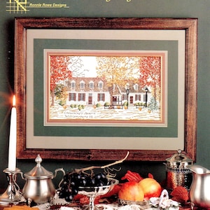 May include: A framed cross-stitch pattern of Chowning's Tavern in Williamsburg, Virginia. The tavern is depicted in autumn colors with trees and a brick building. The image is surrounded by a brown wooden frame and a green mat.