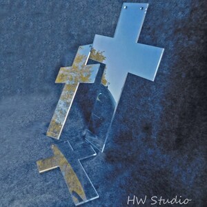 May include: Three clear acrylic crosses of varying sizes are arranged on a textured blue surface. The crosses reflect the surrounding environment, including trees and sky. The image is labeled "HW Studio" in the bottom right corner.