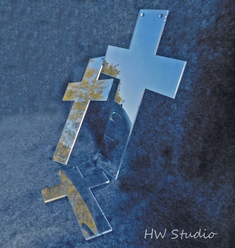 May include: Three clear acrylic crosses of varying sizes are arranged on a textured blue surface. The crosses reflect the surrounding environment, including trees and sky. The image is labeled "HW Studio" in the bottom right corner.