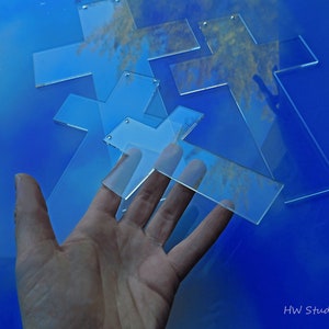 May include: Clear acrylic crosses with small holes for hanging are scattered on a blue surface. The crosses are in various sizes and shapes. The image is well-lit, showing the transparency of the material. The text "HW Studio" is in the bottom right corner.