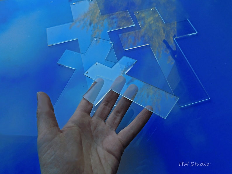 May include: Clear acrylic crosses with small holes for hanging are scattered on a blue surface. The crosses are in various sizes and shapes. The image is well-lit, showing the transparency of the material. The text "HW Studio" is in the bottom right corner.