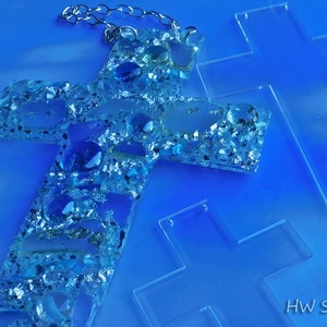 May include: A handcrafted cross pendant with a silver chain, adorned with blue and clear stones and glitter. Two additional clear crosses are also visible. The background is a gradient of blue hues.
