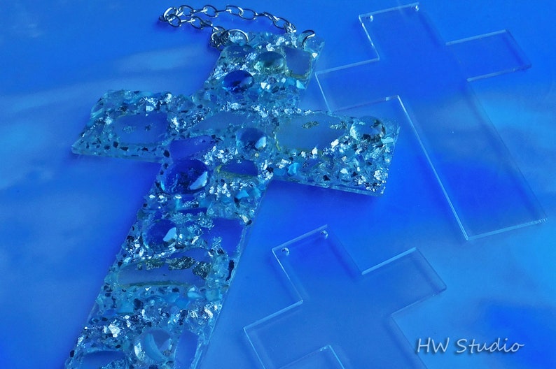 May include: A handcrafted cross pendant with a silver chain, adorned with blue and clear stones and glitter. Two additional clear crosses are also visible. The background is a gradient of blue hues.