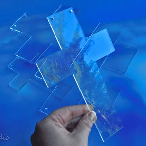 May include: Clear acrylic crosses with rounded corners are scattered on a blue surface. One cross is being held by a hand. The crosses have small holes at the top. The image has a soft focus and a reflective quality.