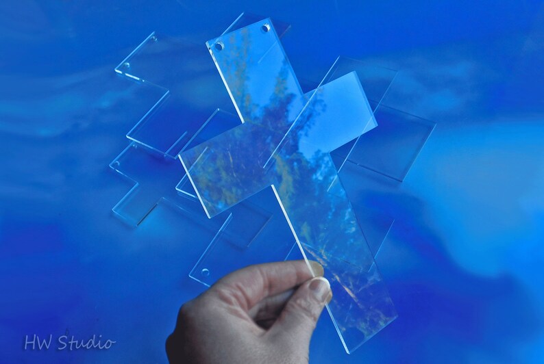 May include: Clear acrylic crosses with rounded corners are scattered on a blue surface. One cross is being held by a hand. The crosses have small holes at the top. The image has a soft focus and a reflective quality.