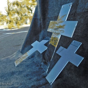 May include: Three clear acrylic crosses with reflective surfaces are arranged on a dark gray, textured background. The crosses vary in size and are positioned at different angles, creating a dynamic composition. The crosses have small holes for hanging.