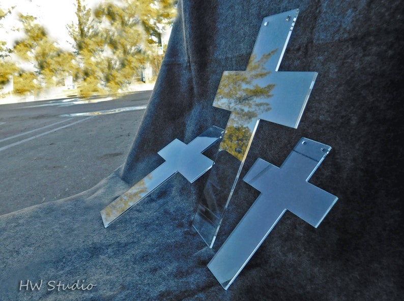 May include: Three clear acrylic crosses with reflective surfaces are arranged on a dark gray, textured background. The crosses vary in size and are positioned at different angles, creating a dynamic composition. The crosses have small holes for hanging.