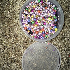 Pode incluir: Um recipiente de plástico transparente cheio de slime colorido com contas de arco-íris. O slime está em um recipiente de plástico transparente com tampa. O recipiente está em um tapete marrom.