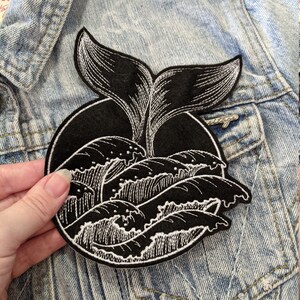 May include: Black and white embroidered patch of a whale tail breaching out of the water. The patch is circular and has a black felt background.