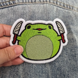 May include: A green frog with a red mouth holding two white knives with red handles. The frog is embroidered on a white patch.