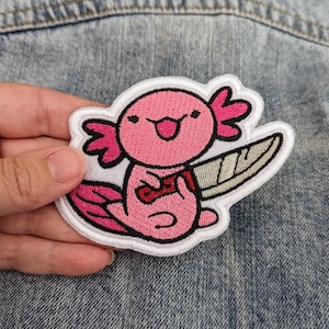 May include: A pink axolotl with a knife embroidered patch. The axolotl is smiling and holding the knife in its front paws.