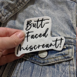 Peut inclure: Écusson thermocollant blanc avec du texte brodé noir qui dit "Butt-Faced Miscreant!" L'écusson est sur une veste en jean bleu clair.