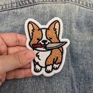May include: A white, brown, and red embroidered patch of a corgi dog holding a knife in its mouth. The patch is on a blue denim background.