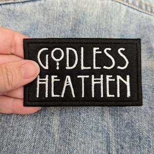 May include: A black rectangular patch with white embroidered text that reads "GODLESS HEATHEN". The patch is held against a light blue denim jacket.